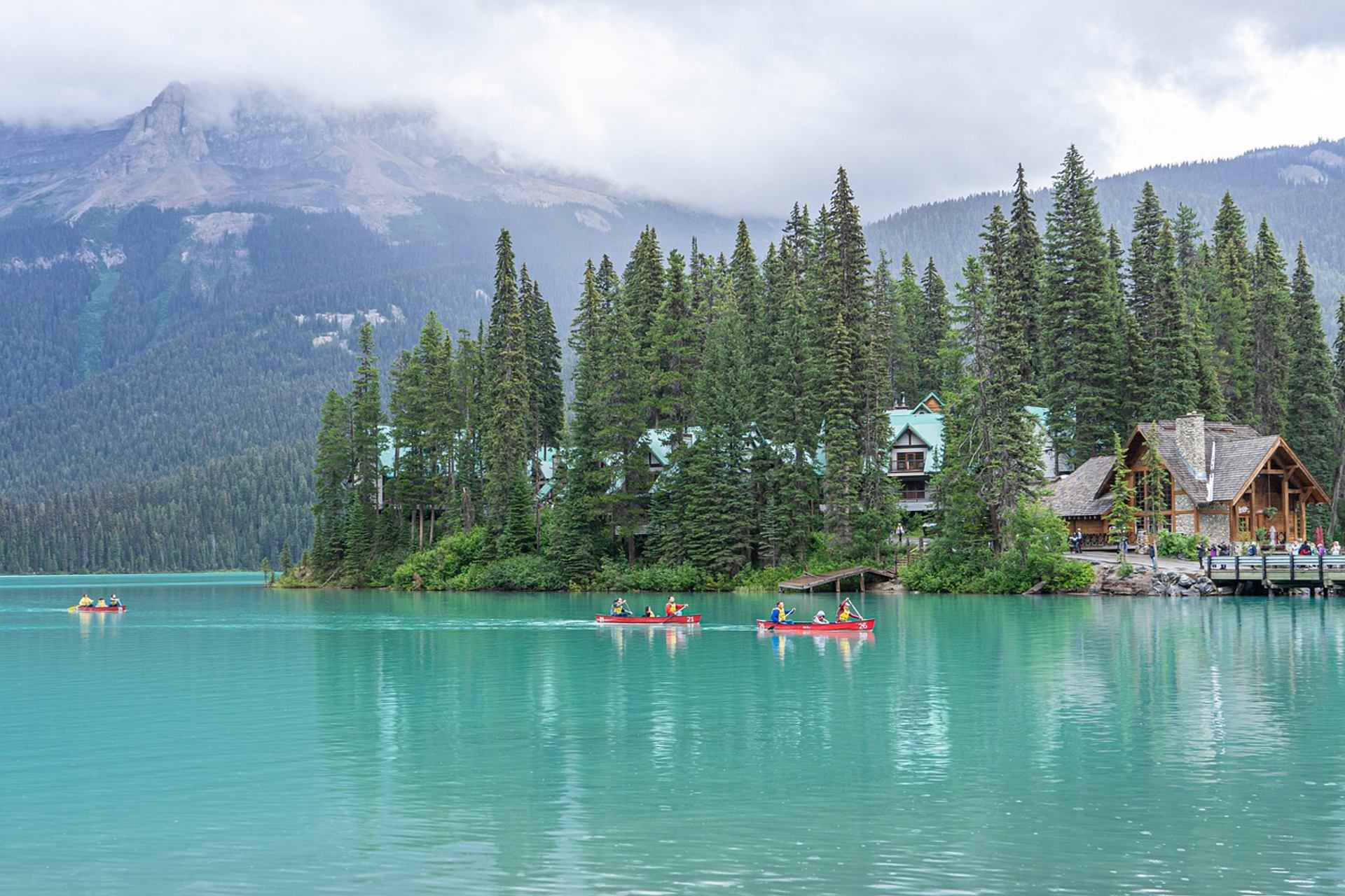 Turquoise lake with canoes, forested shoreline, cabins and misty mountains in background.
