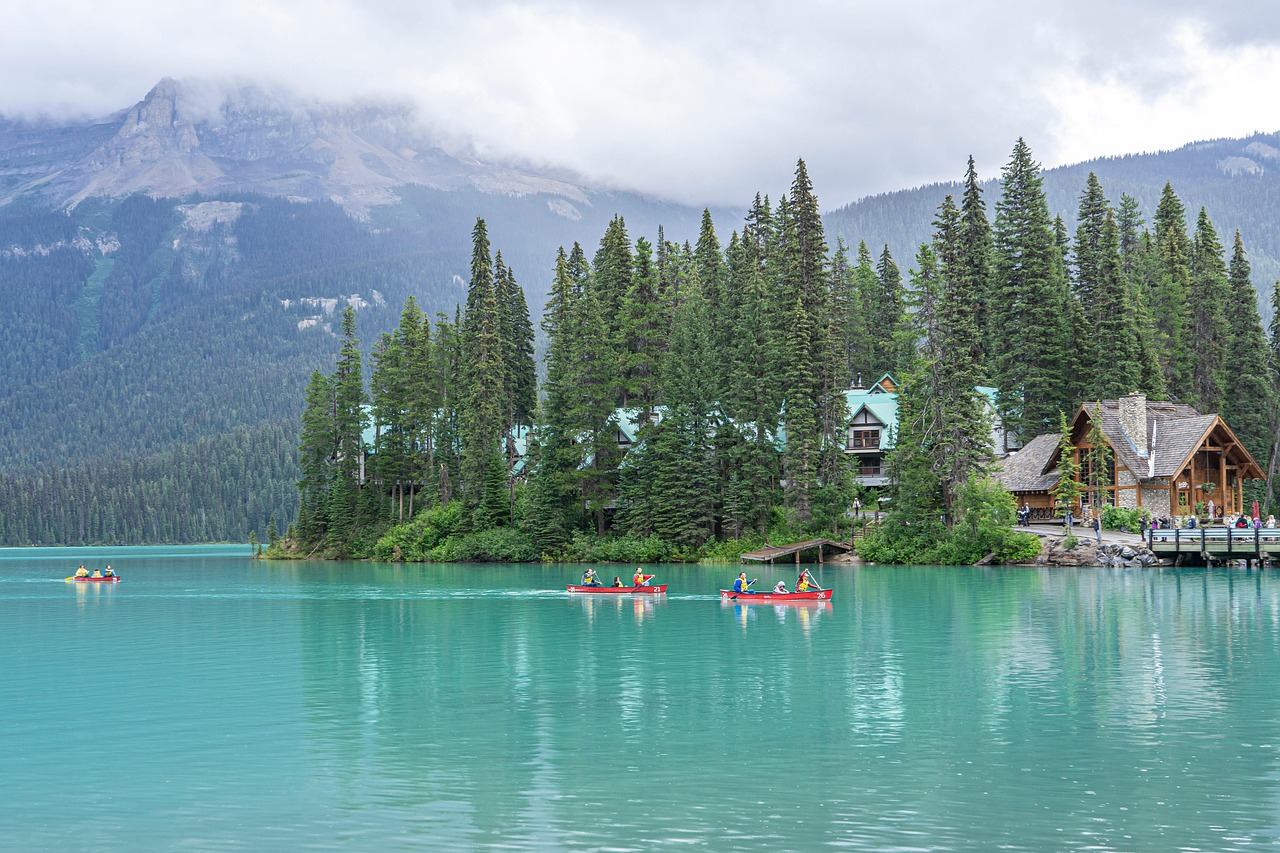 Turquoise lake with canoes, forested shoreline, cabins and misty mountains in background.