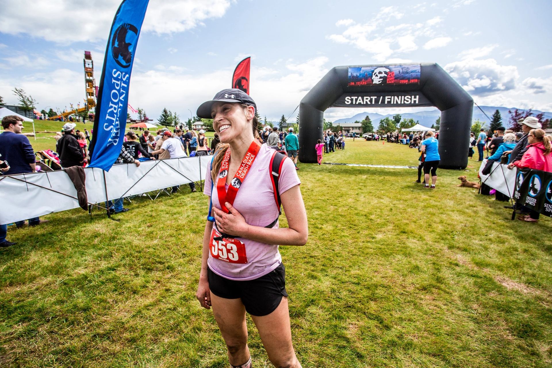 Runner with medal stands near the Canadian Death Race finish arch on grassy field.