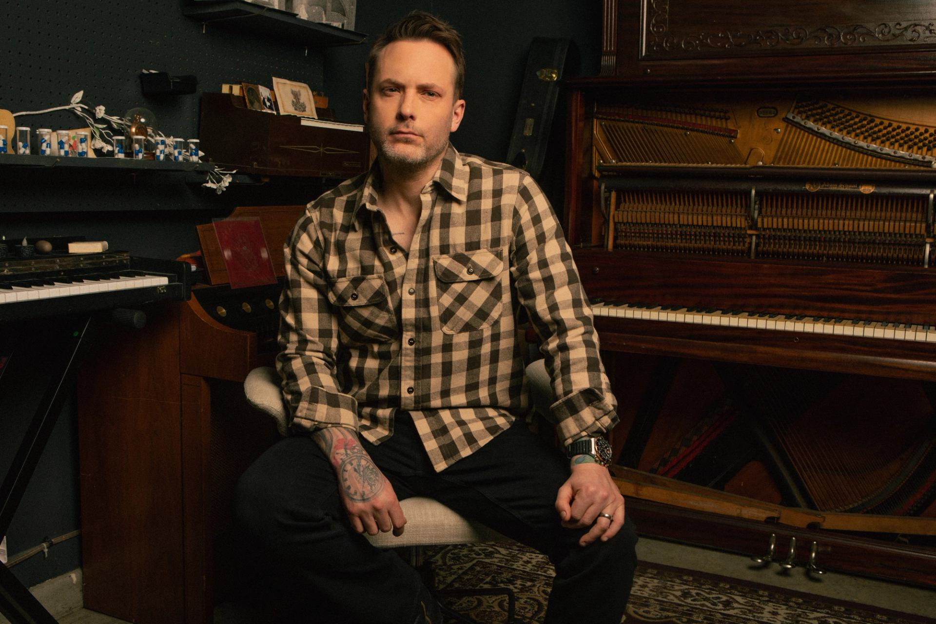A man with tattoos and a plaid shirt sits in a room with a keyboard and an open upright piano.