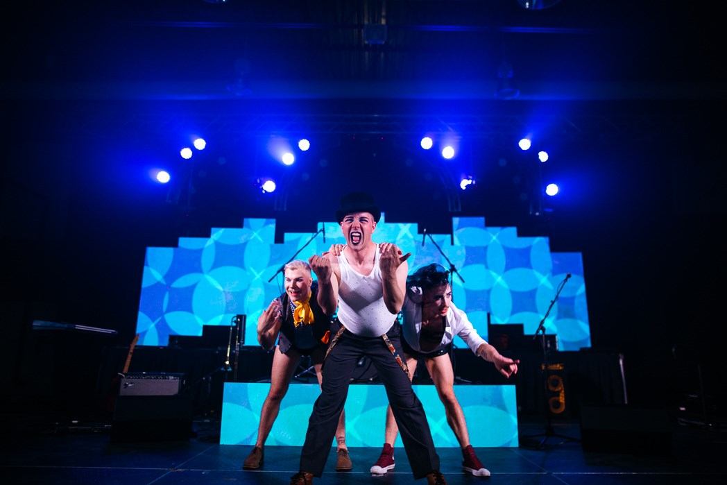 Three performers dancing on a brightly lit stage with blue lights behind them.
