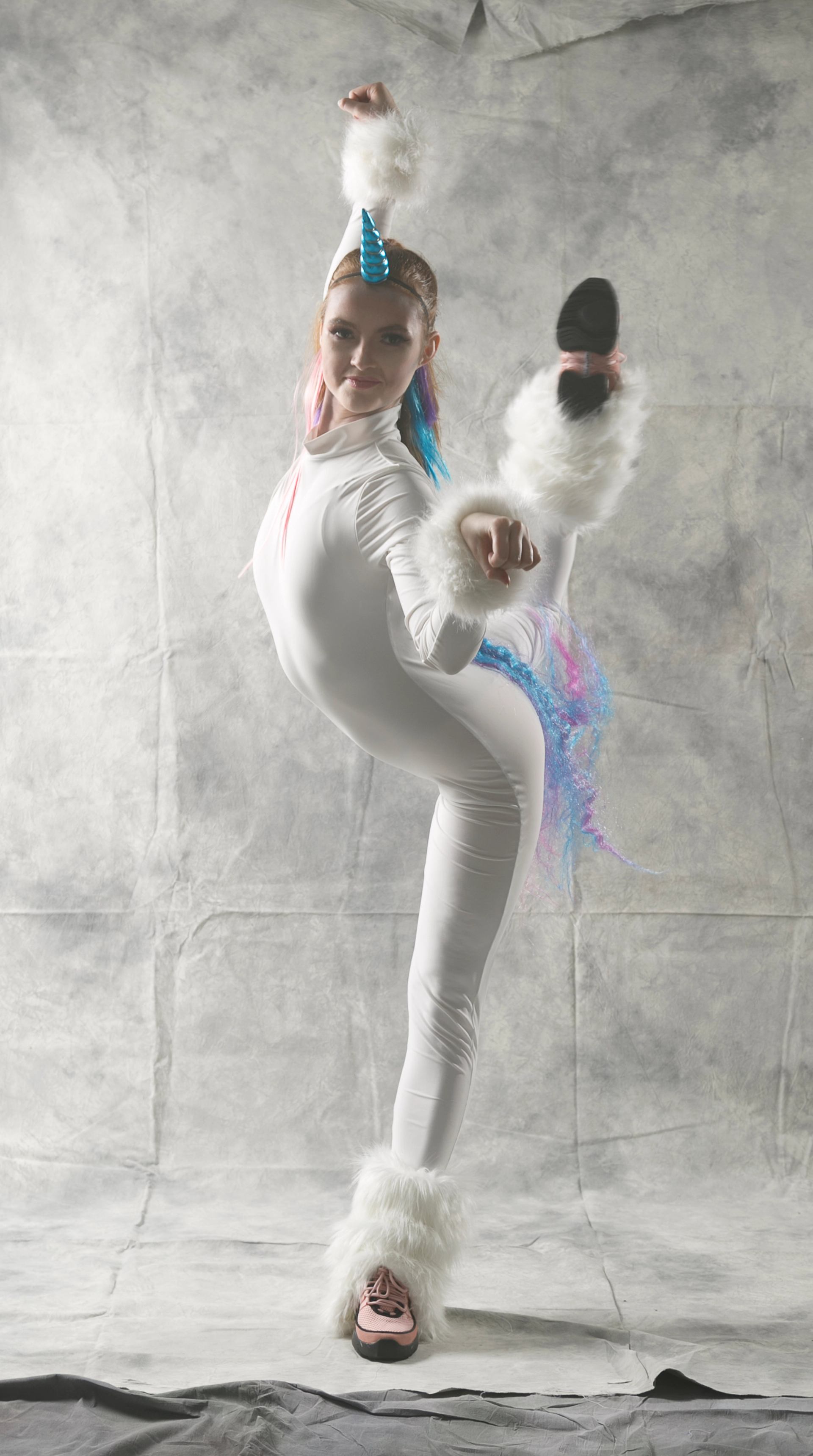Dancer in white unicorn costume performs high kick against plain studio backdrop.