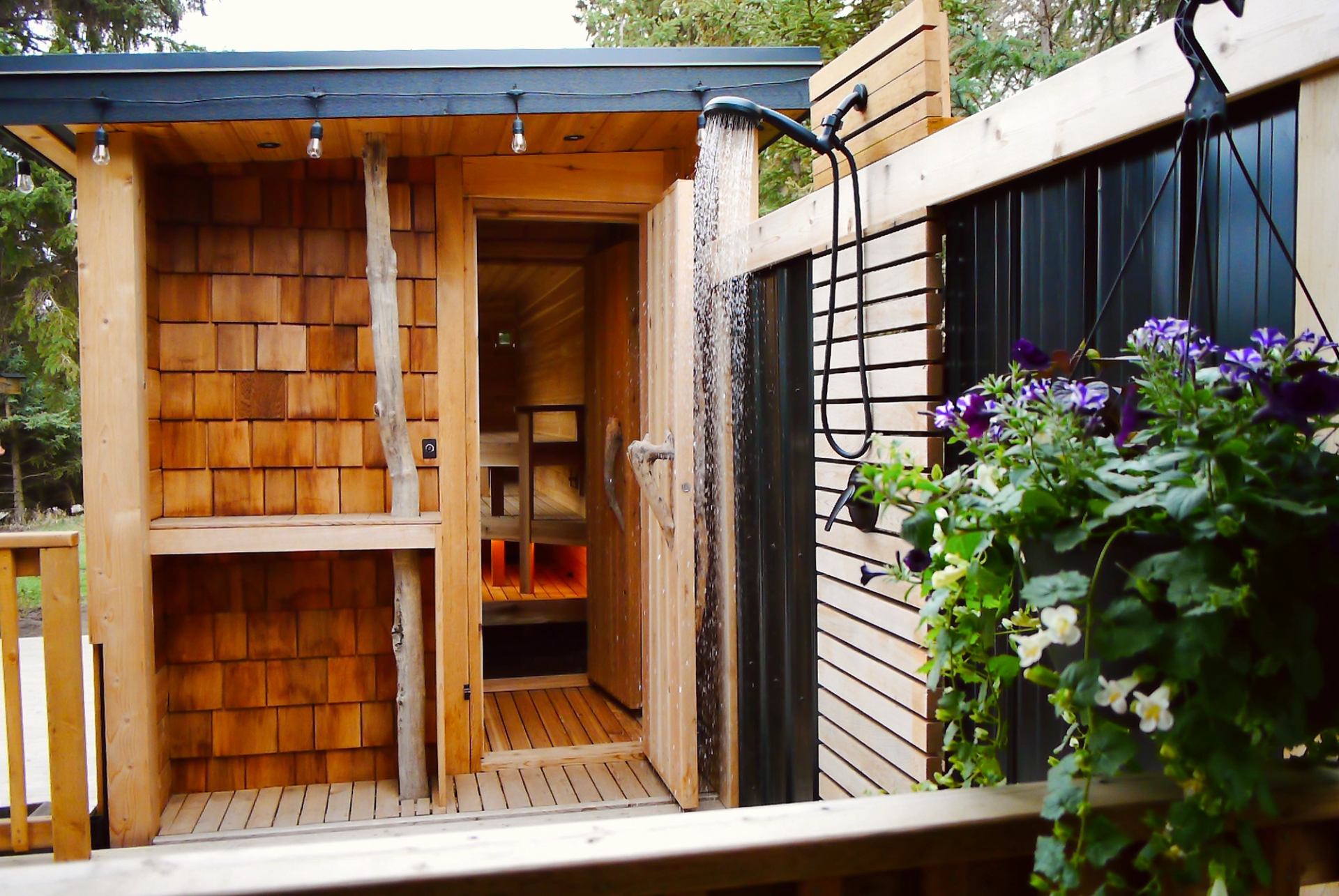  A wooden outdoor sauna entrance opens onto a small deck with a mounted shower pouring water beside it. Warm wood textures, string lights, and greenery create a peaceful, spa-like setting.