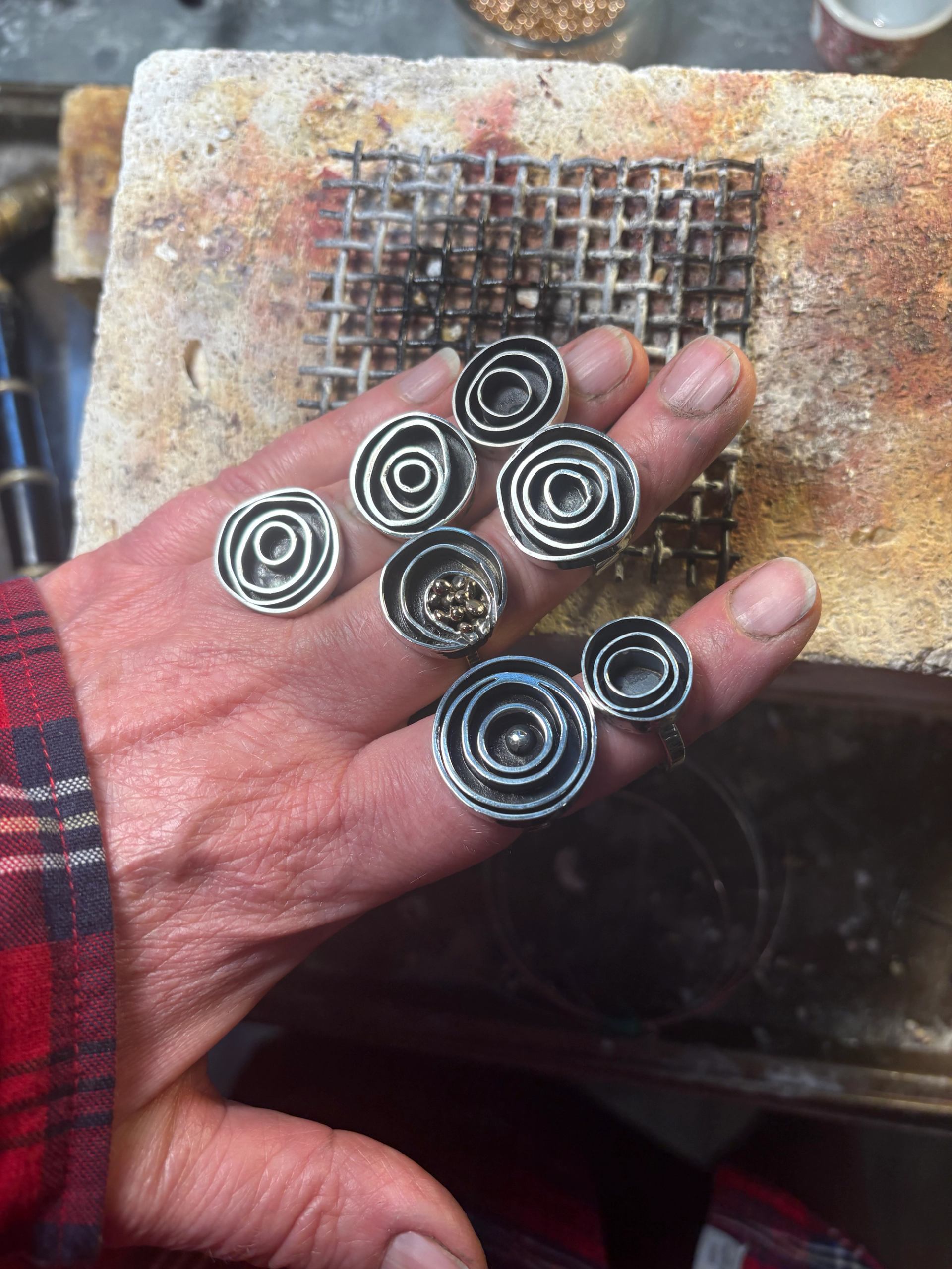 Hand wearing multiple spiral metal rings above a soldering block and wire mesh.