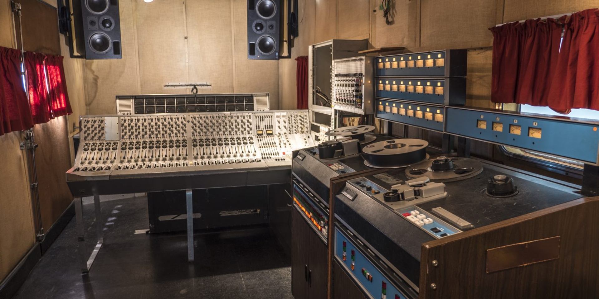 Vintage recording studio with large mixing console, reel-to-reel machines and wall meters.