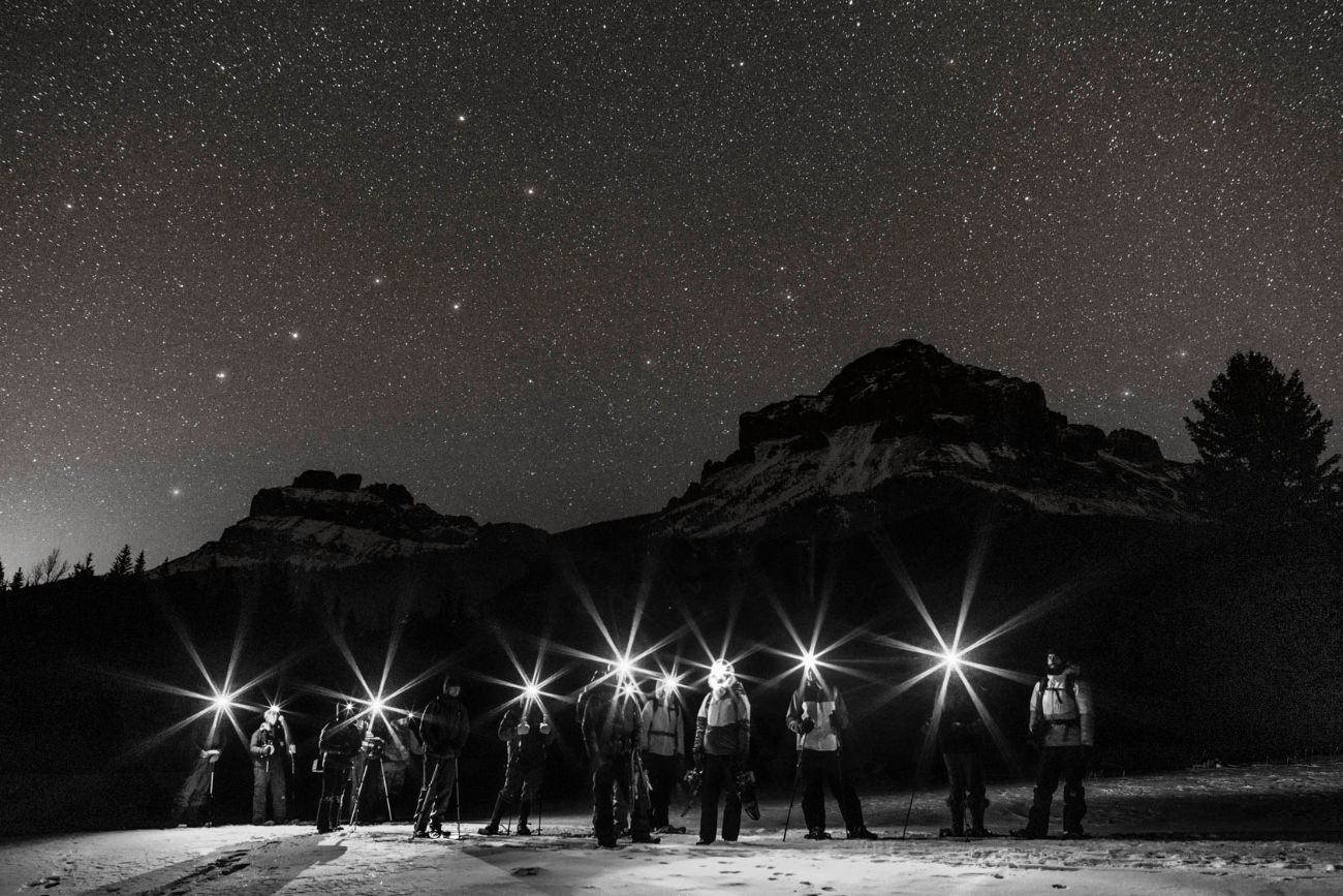 Black and white photo of a group with bright headlamps under a starry night sky and mountains.