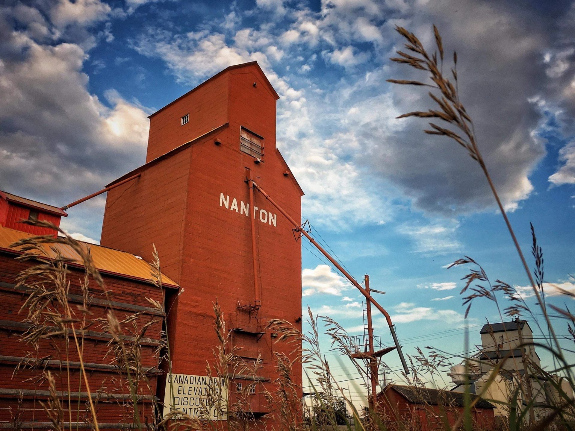A red grain elevator beneath a sky of swirling clouds with grain in the foreground.