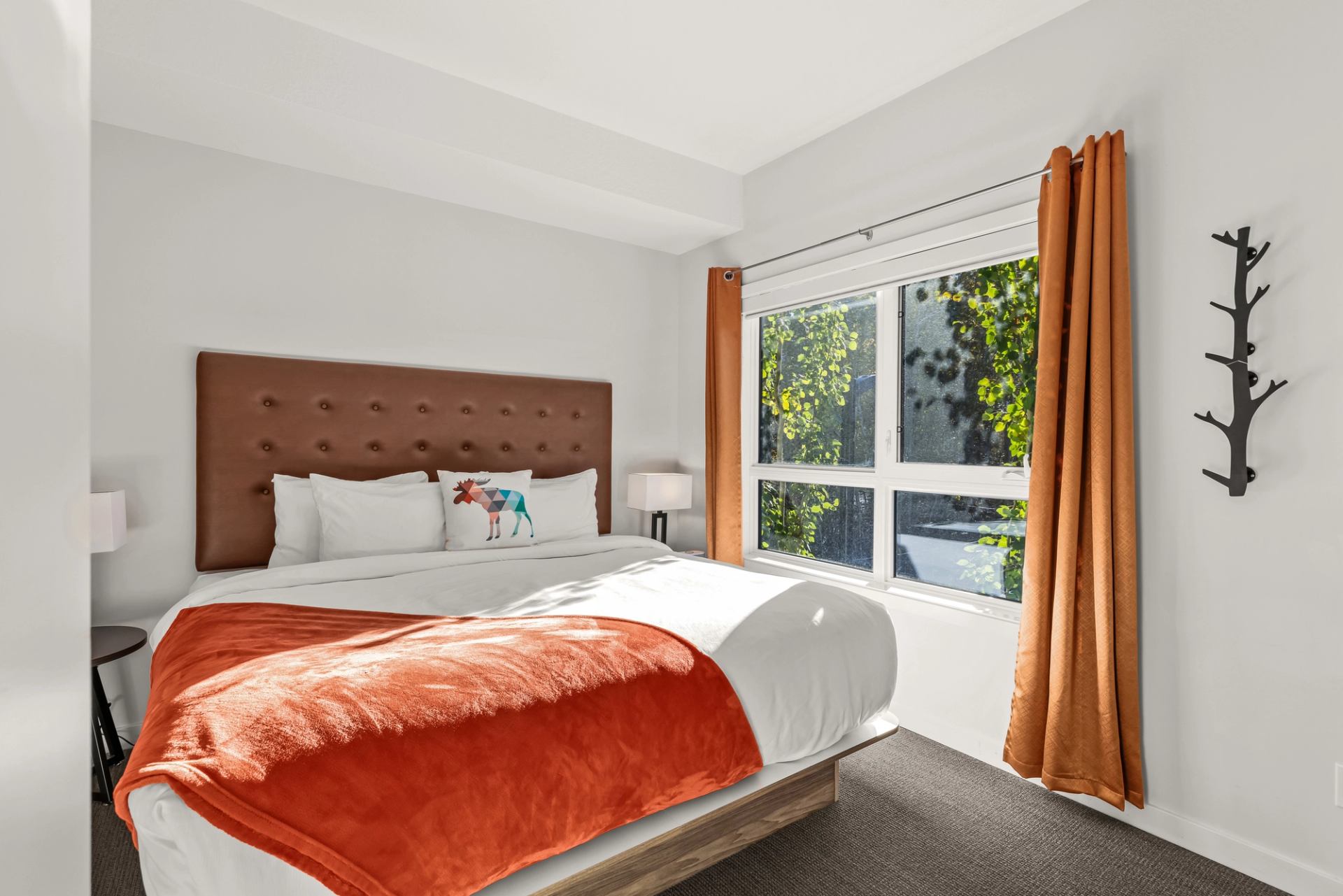 Bedroom with brown headboard, orange blanket and curtains beside a large window.