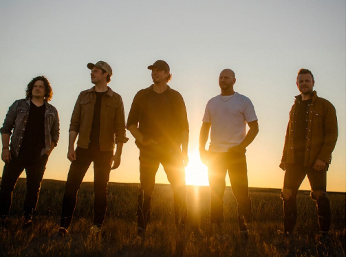 Five people stand in a field with the sun low on the horizon behind them.