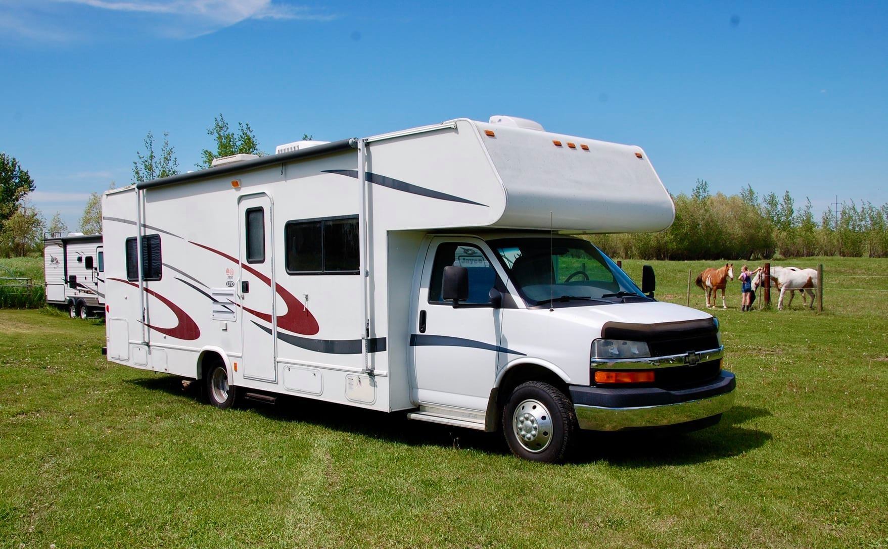 White RV on grassy field with trees, horses, and people in the background.