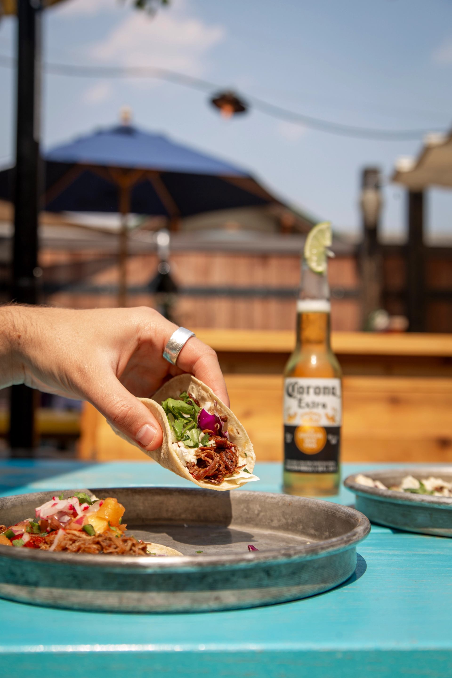 A hand holds a taco with meat and fresh toppings over a blue table, with a blurred Corona beer bottle and outdoor patio in the background.