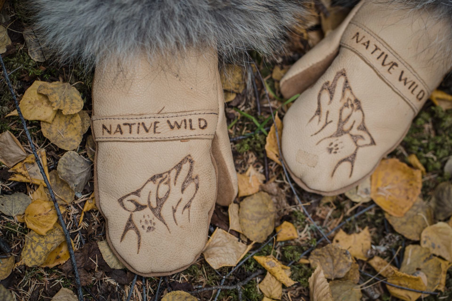 Leather mittens with wolf art and "Native Wild" text, lying on autumn leaves and forest ground.