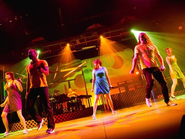 Five performers on stage with colorful lighting and live band in the background.
