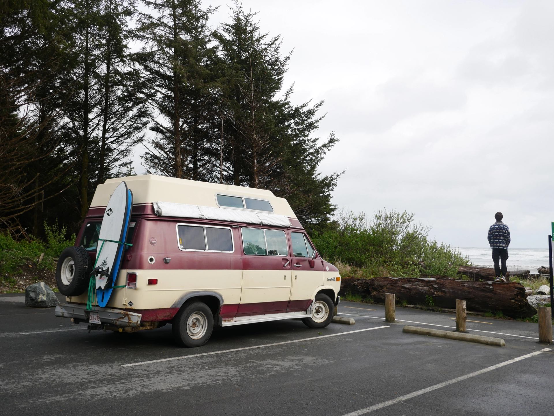 Maroon camper van with surfboard in forested coastal parking lot.