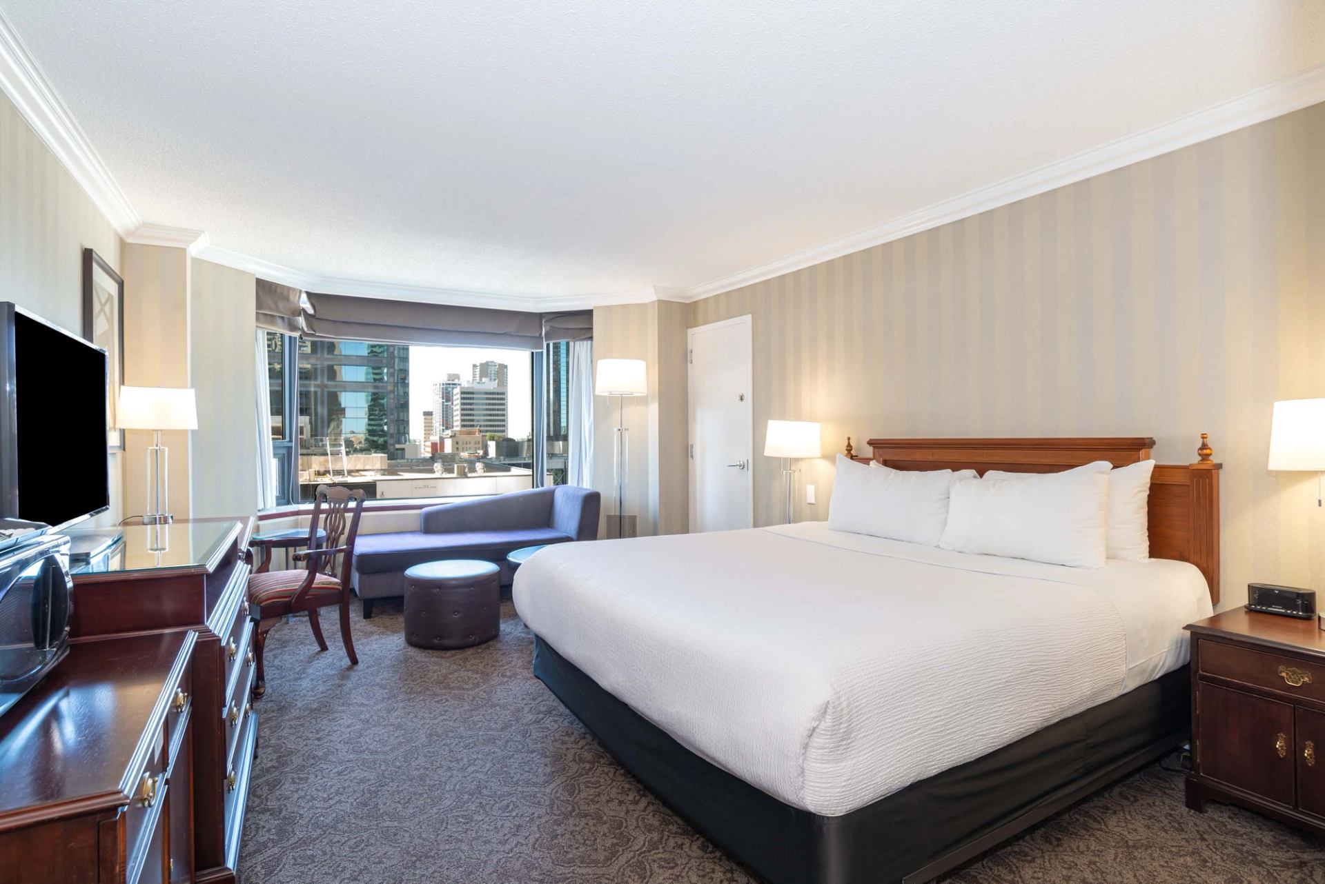Hotel room with king bed, sofa, desk, and city view through large window.