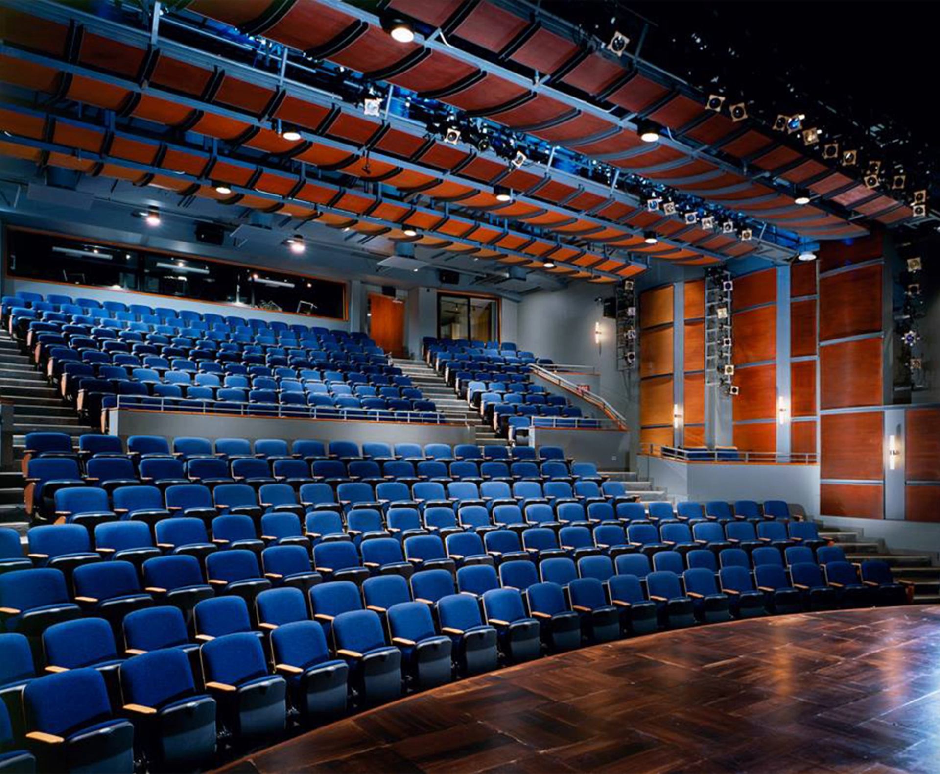 Empty modern theatre auditorium with blue seats, wooden stage and overhead lighting fixtures.