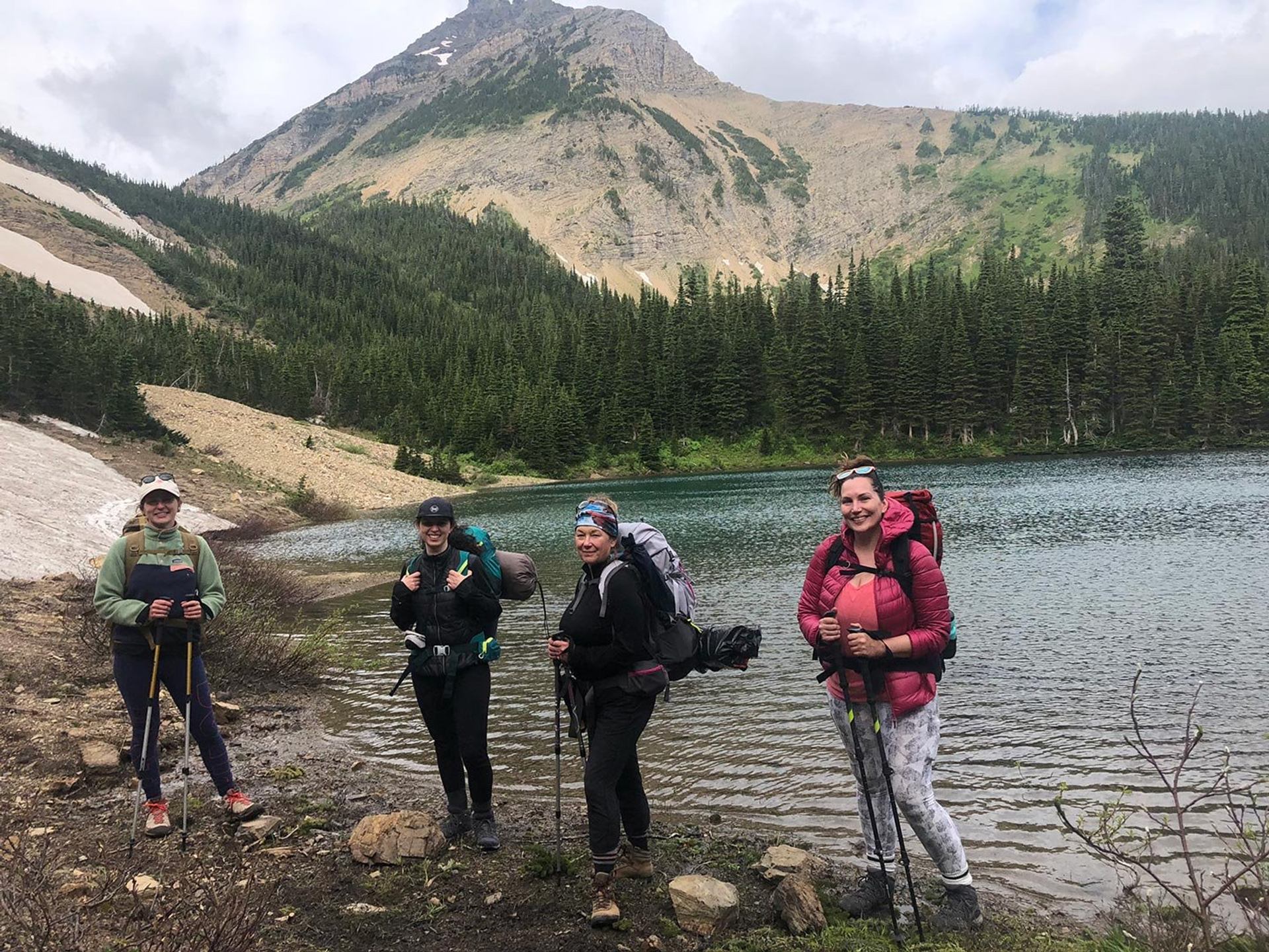 Hikers with backpacks stand by a mountain lake, forested slopes rising behind them.