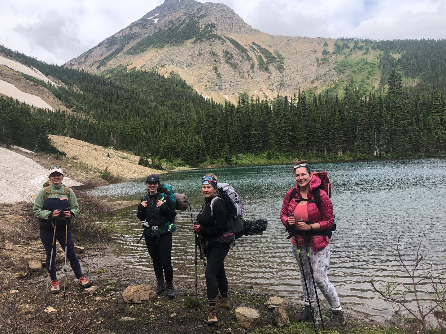 Hikers with backpacks stand by a mountain lake, forested slopes rising behind them.