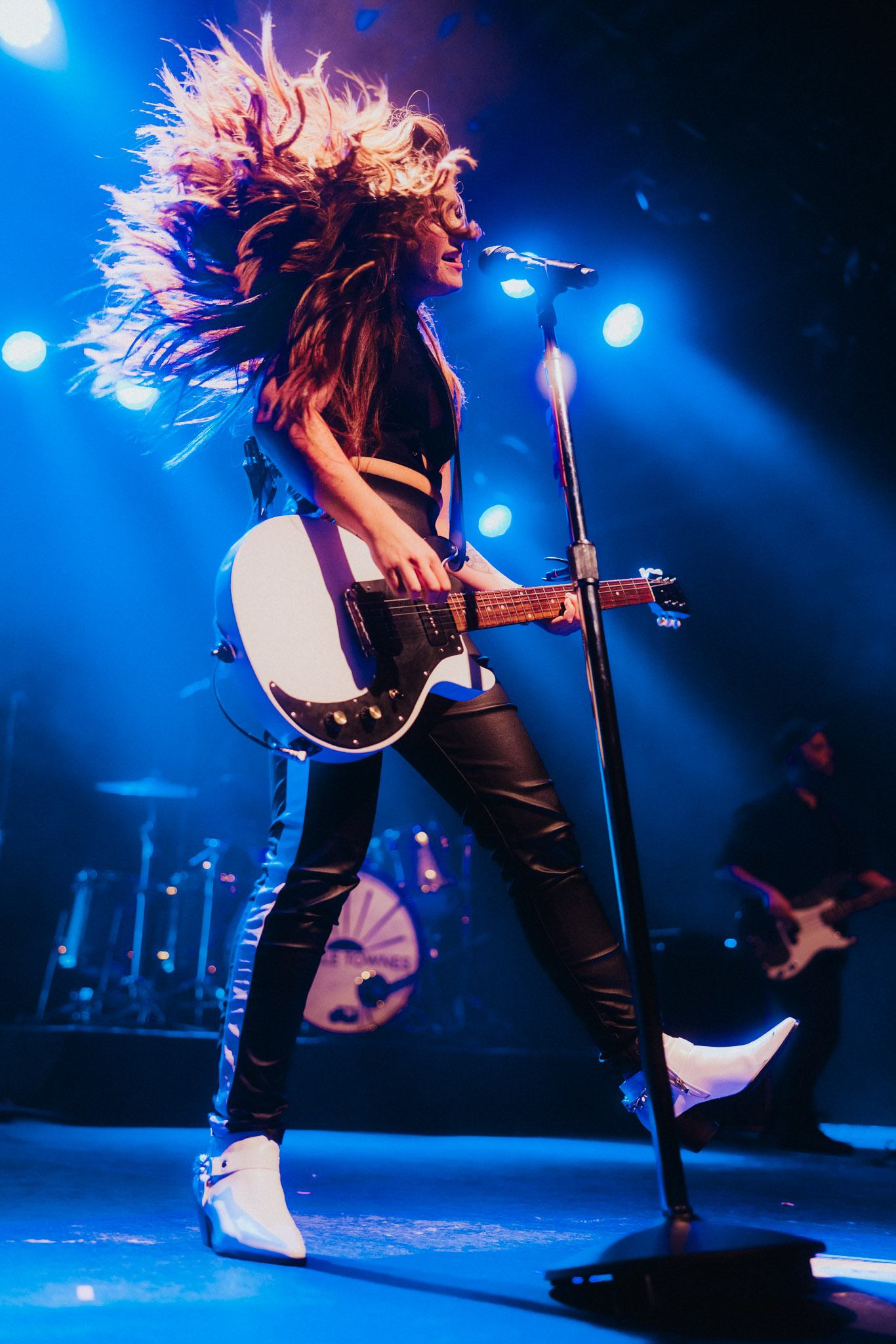 Guitarist with long hair performs on stage under blue lights, microphone and drums behind.