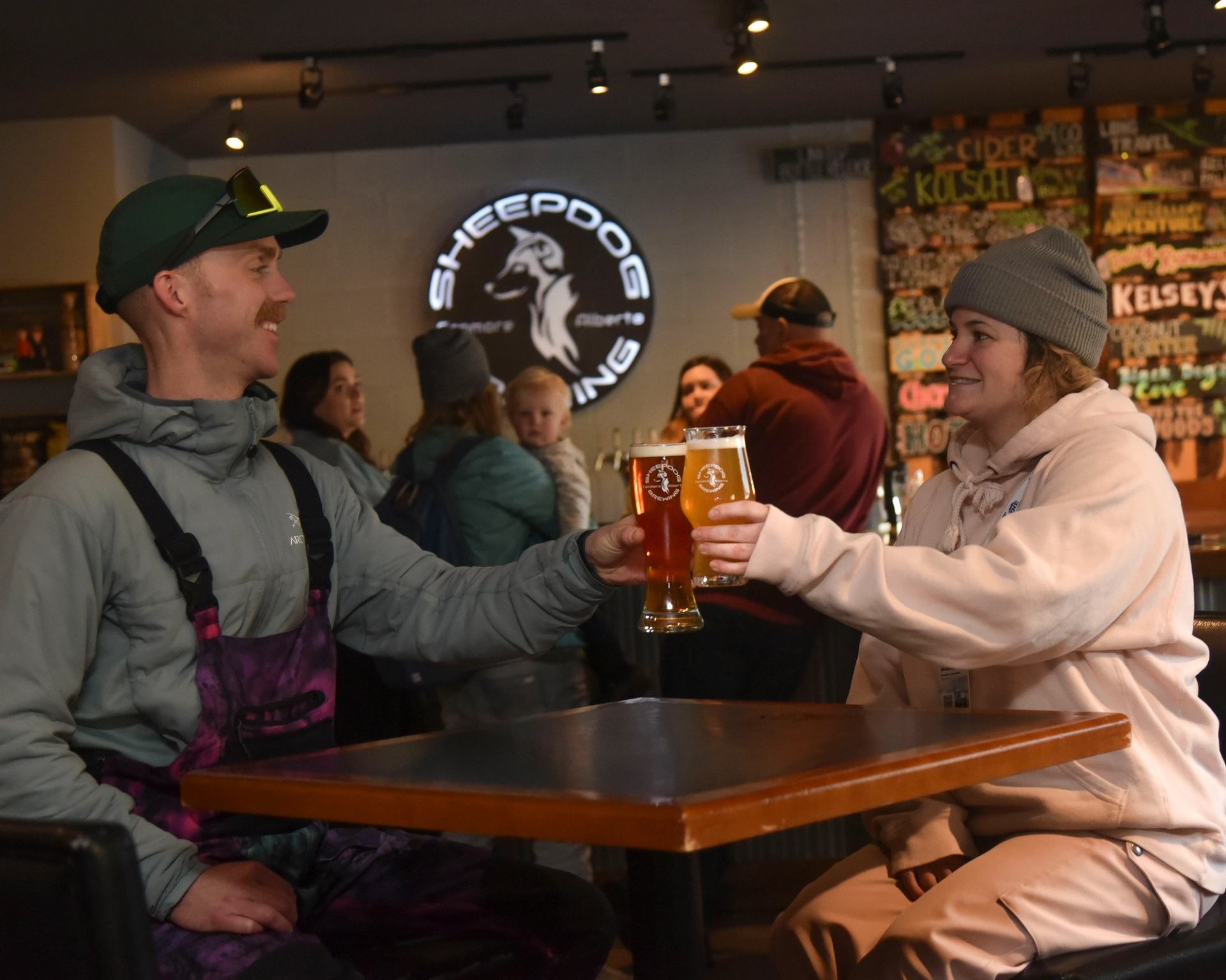 Two people clink glasses of beer at a table inside Sheepdog Brewing.