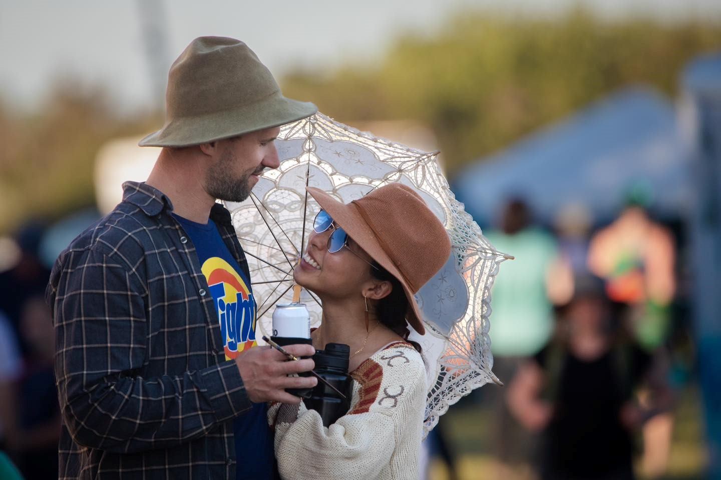 Two festival-goers smile under an umbrella, holding drinks outdoors at South Country Fair.