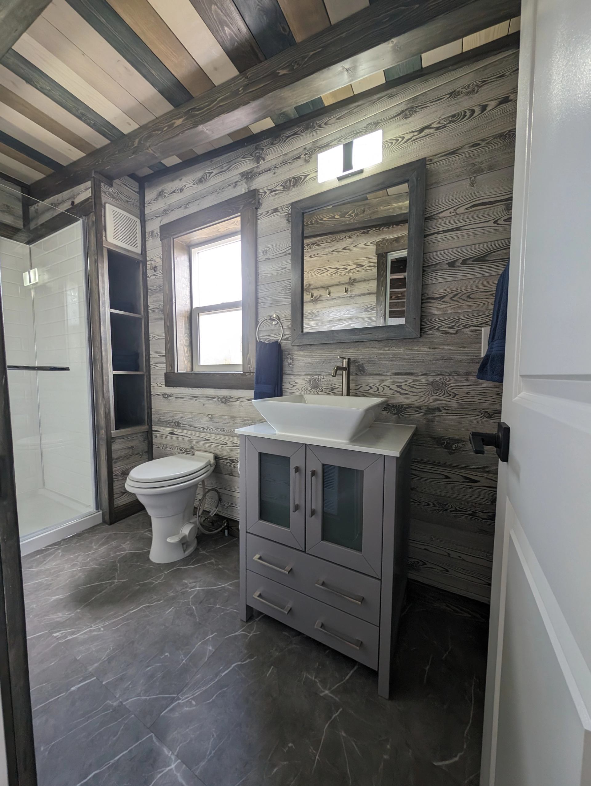 Bathroom with wood-panelled walls, glass shower, toilet, vessel sink and grey vanity.