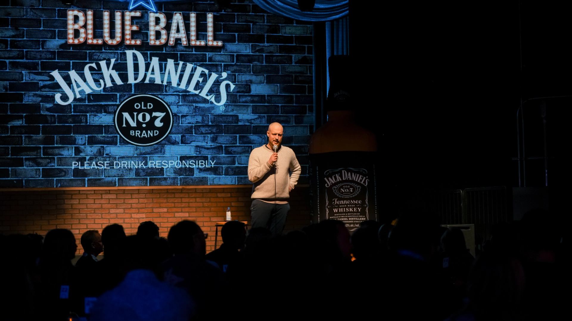 A man performs stand-up comedy on stage with a microphone during a lively show. The backdrop displays "Jack Daniel's" and "Blue Ball".