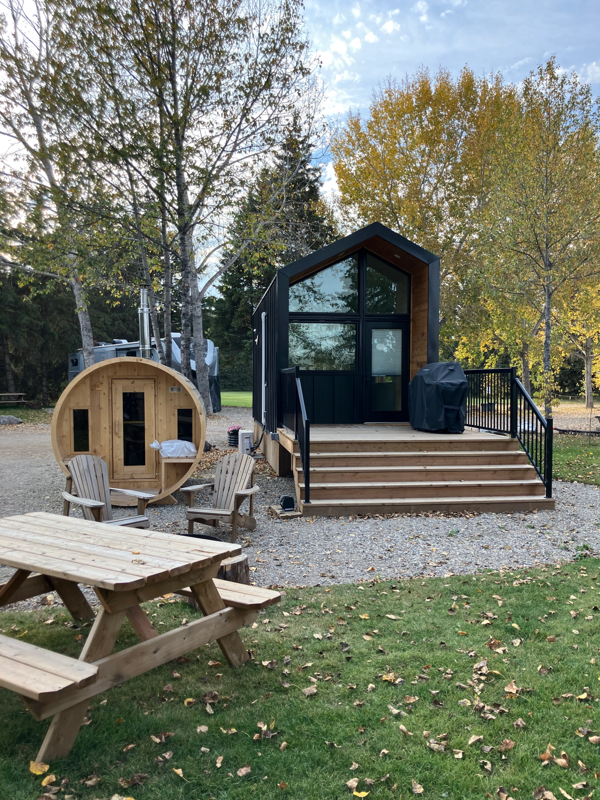 Small modern cabin with steps, a wooden sauna, and a picnic table set among trees with autumn leaves.