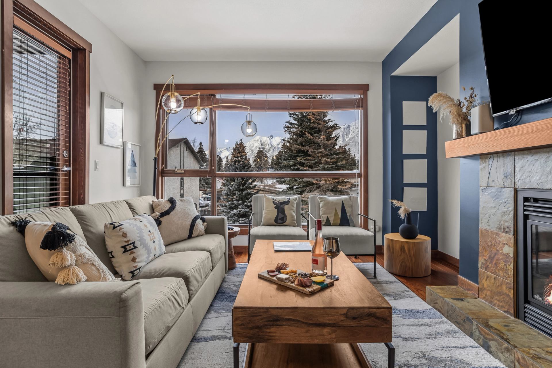 Living room with sofa, fireplace, large window, and mountain view behind evergreen trees.
