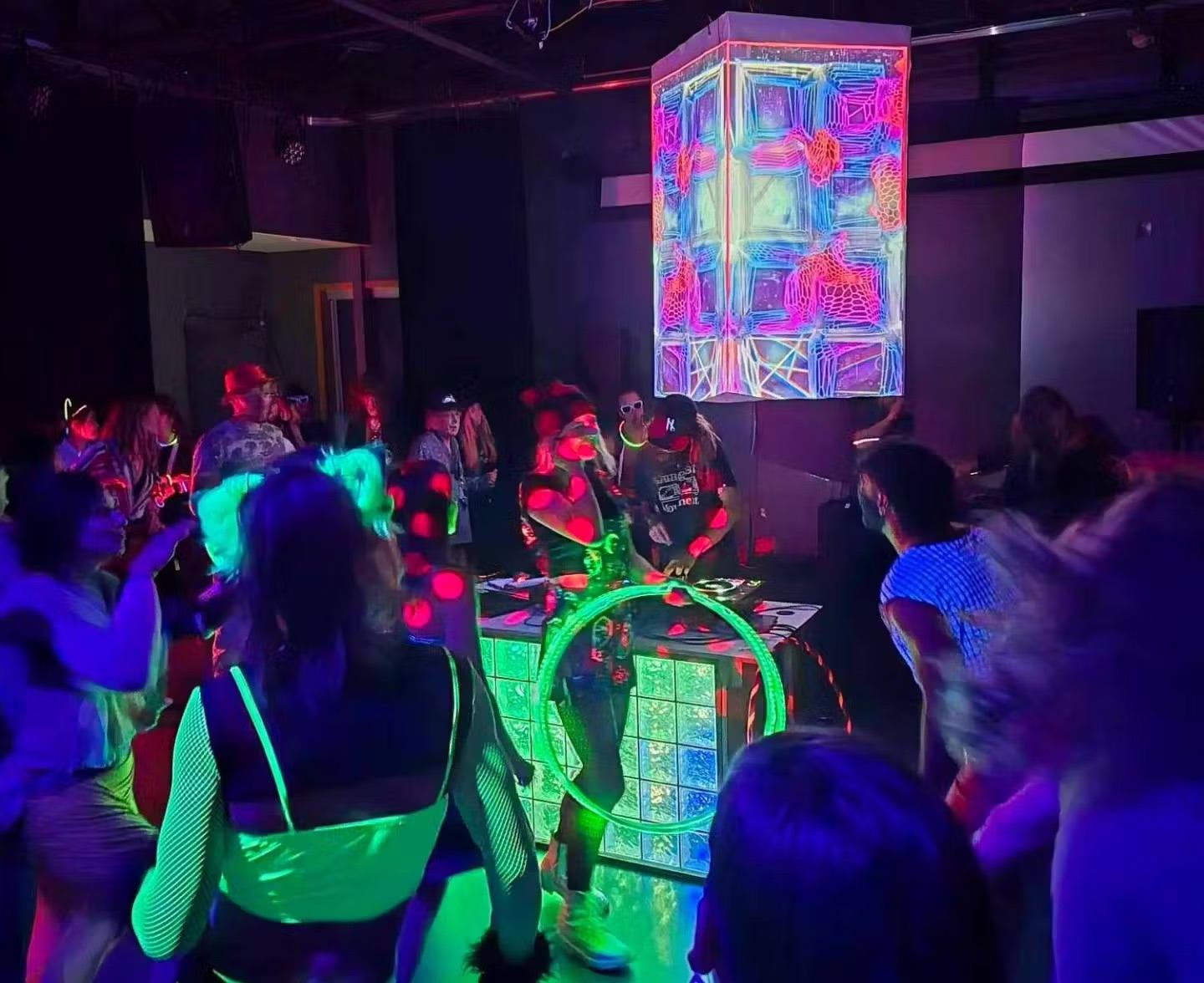 Crowd dances under neon lights at an indoor party with glowing decor and a vibrant DJ setup.