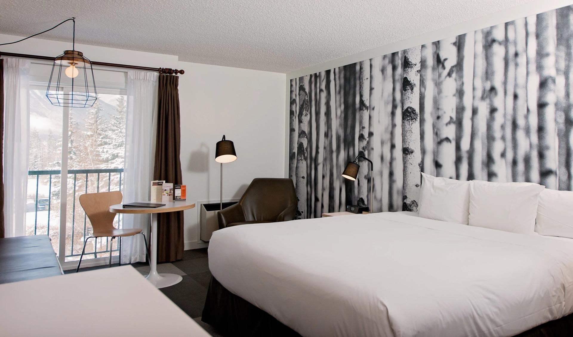 Modern hotel room with birch tree wallpaper, balcony, and cozy seating.