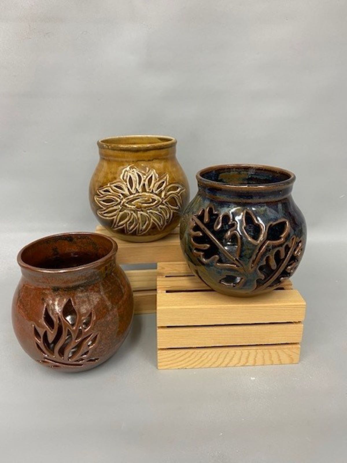 Three carved ceramic vessels with leaf patterns and earthy glazes displayed on wooden blocks.