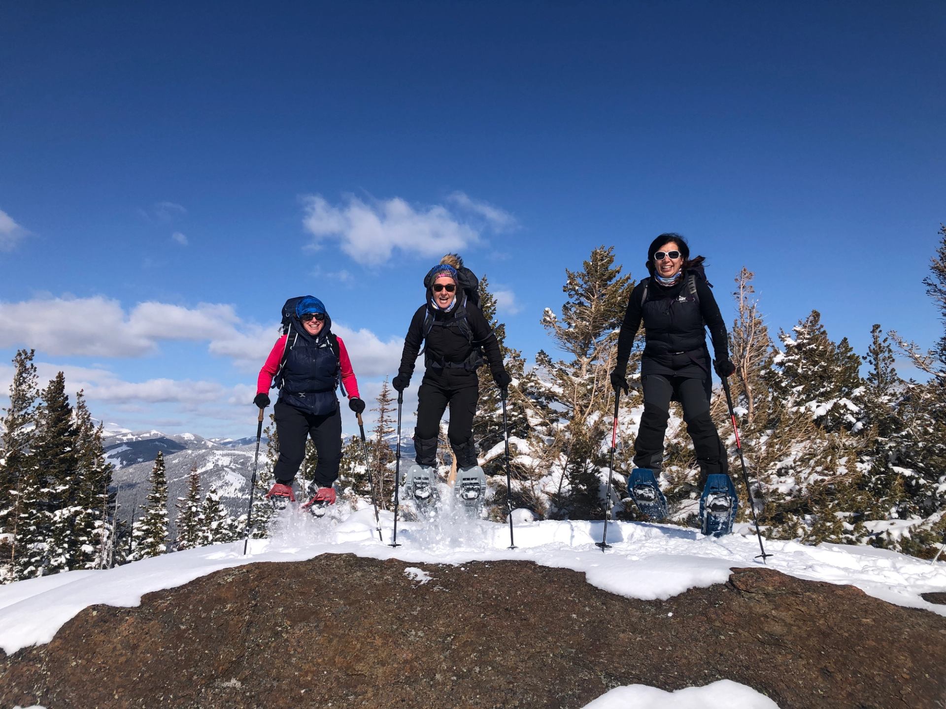 Three snowshoers jumping off a snowy rock with pine trees and blue sky in the background.