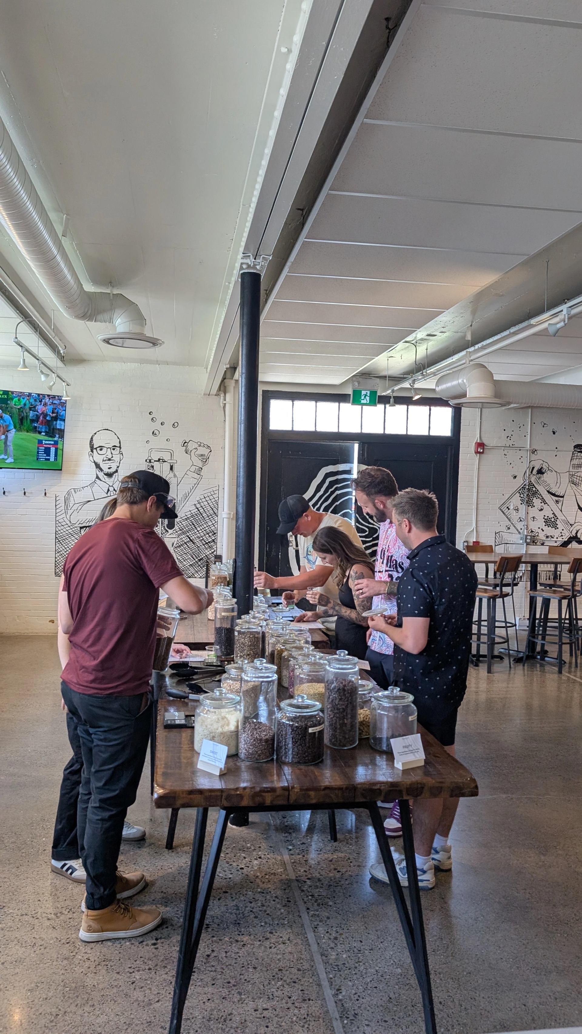 People examine glass jars of ingredients arranged on a long wooden table.