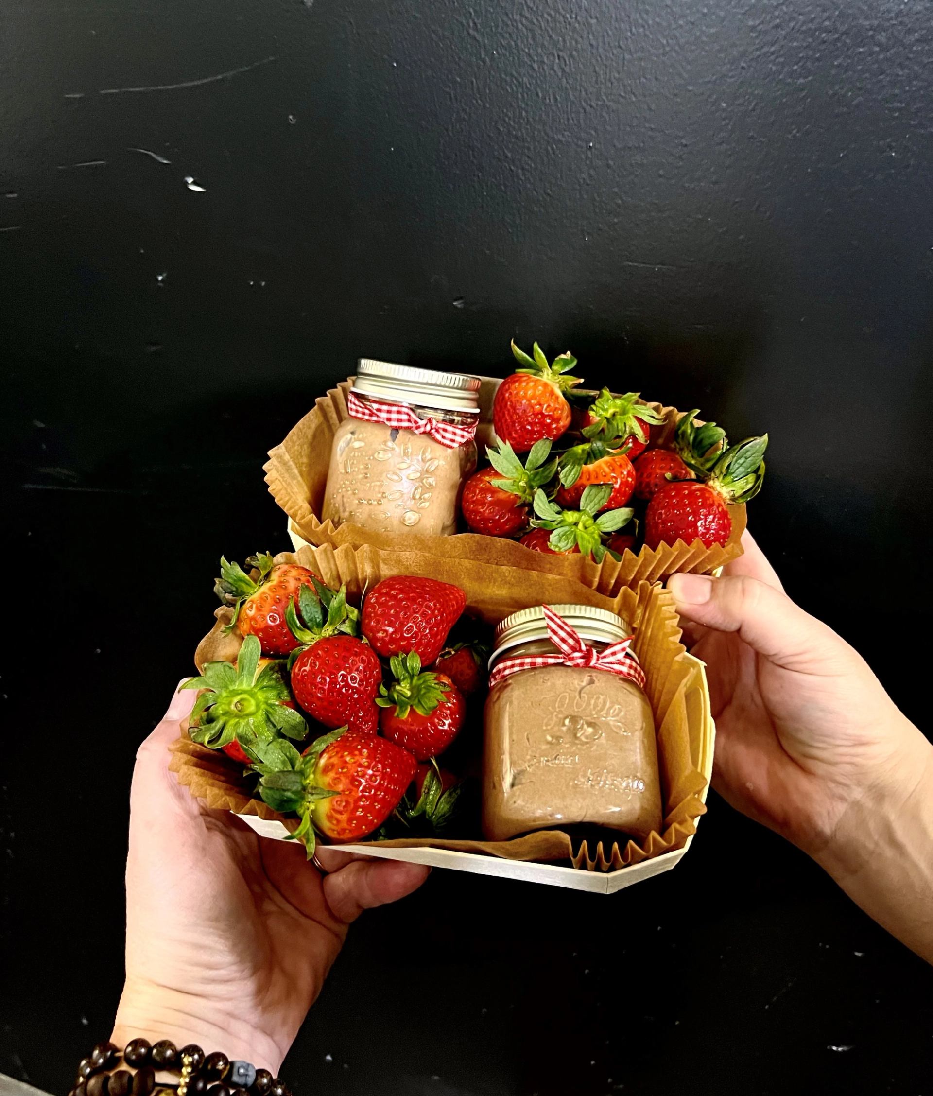 Hand holding a gift basket with strawberries and two glass jars with red gingham lids.