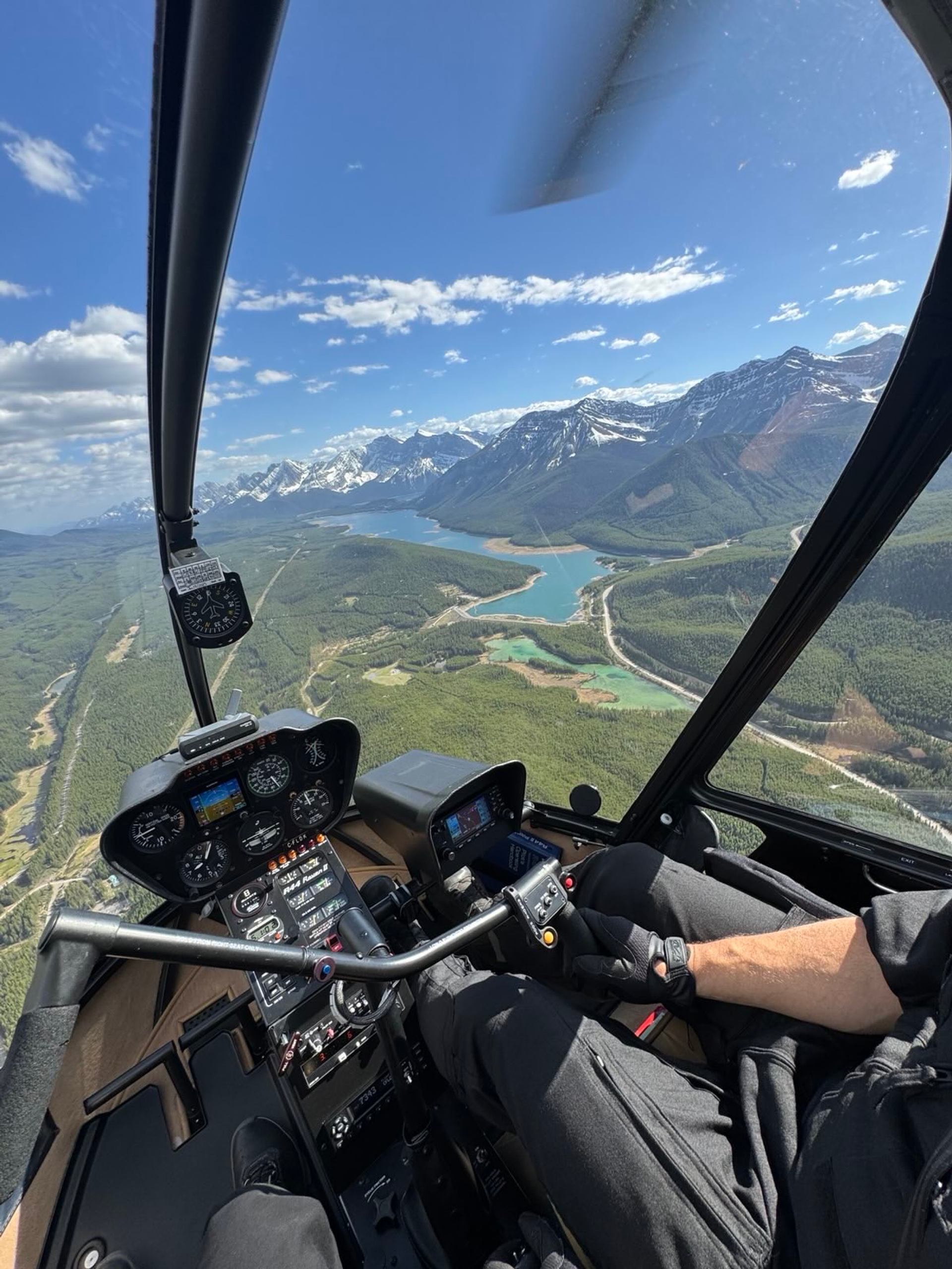 Helicopter cockpit view over forested valley, winding river and distant snow-capped mountains.