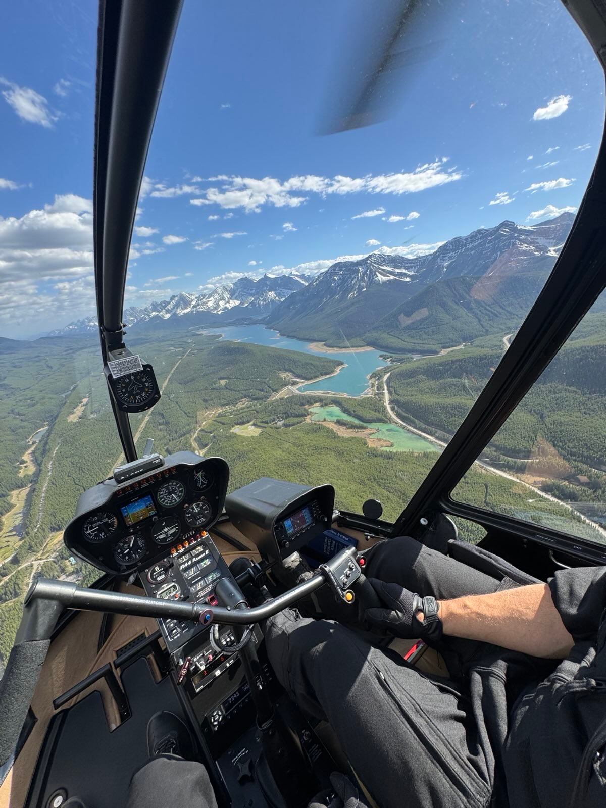 Helicopter cockpit view over forested valley, winding river and distant snow-capped mountains.