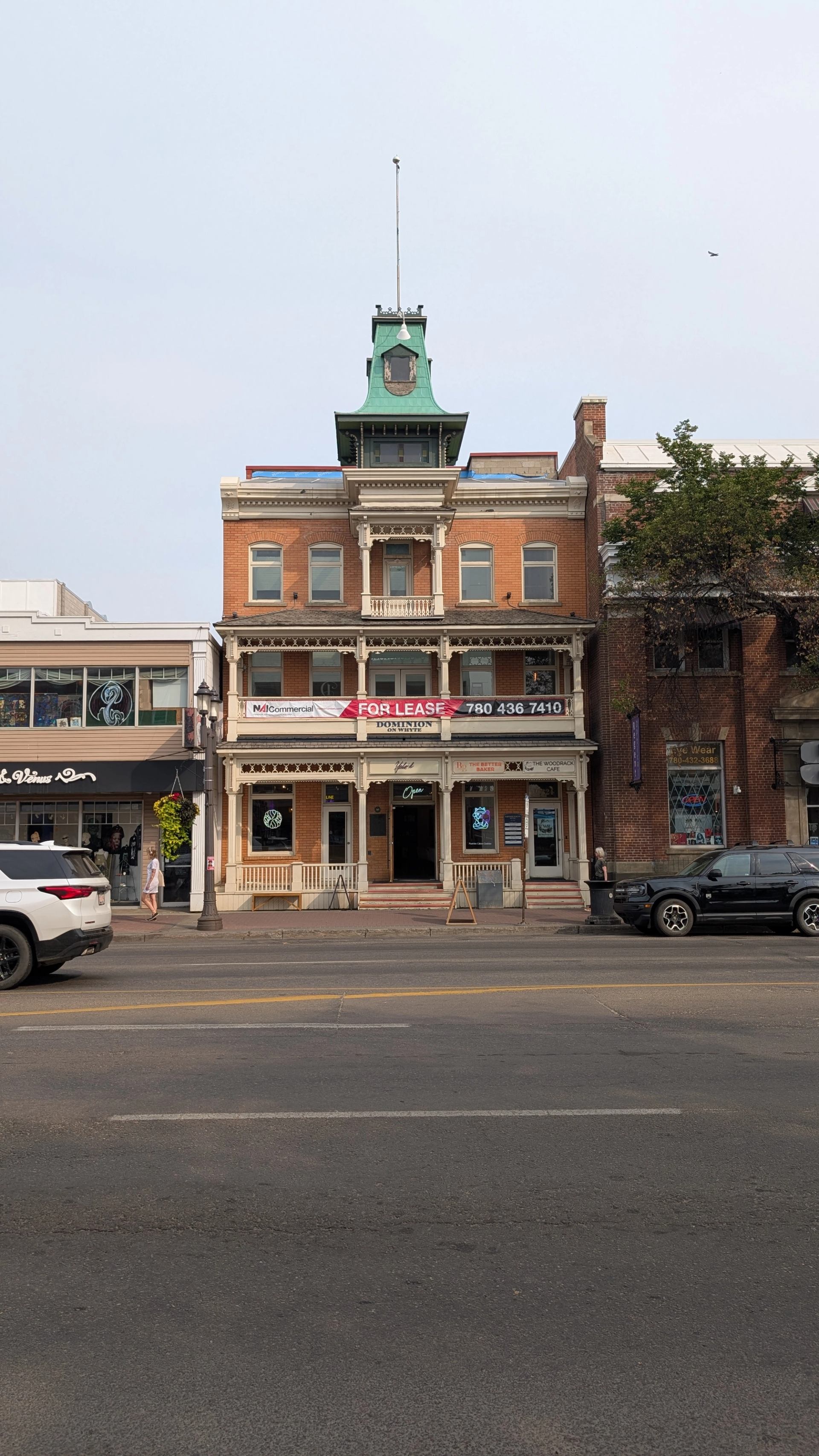 Historic brick building with green tower, for lease banner, shops and parked vehicles.