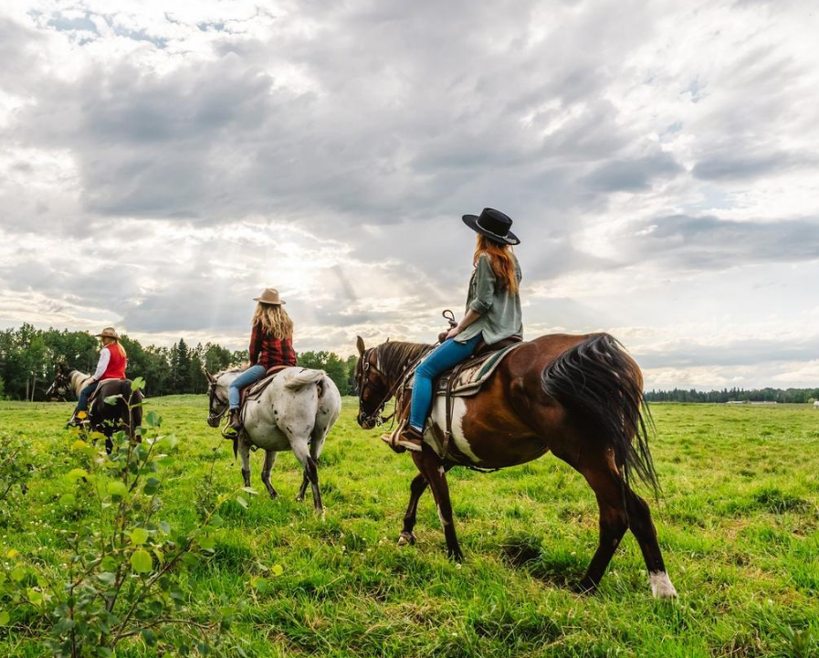 Horseback riders cross a green field beneath a cloudy sky.