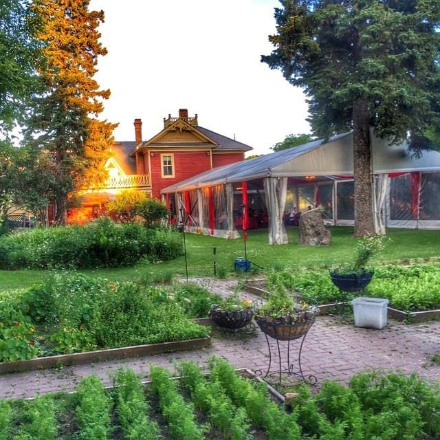 A picturesque scene of a red house and garden, featuring a tented area with glowing lights, surrounded by lush greenery and a stone pathway at sunset.