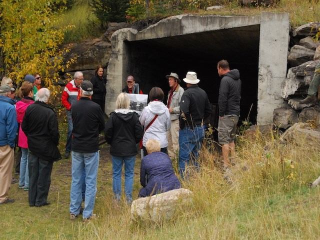 Group gathers outdoors by concrete underpass; guide speaks with visual aid amid autumn trees and grass.