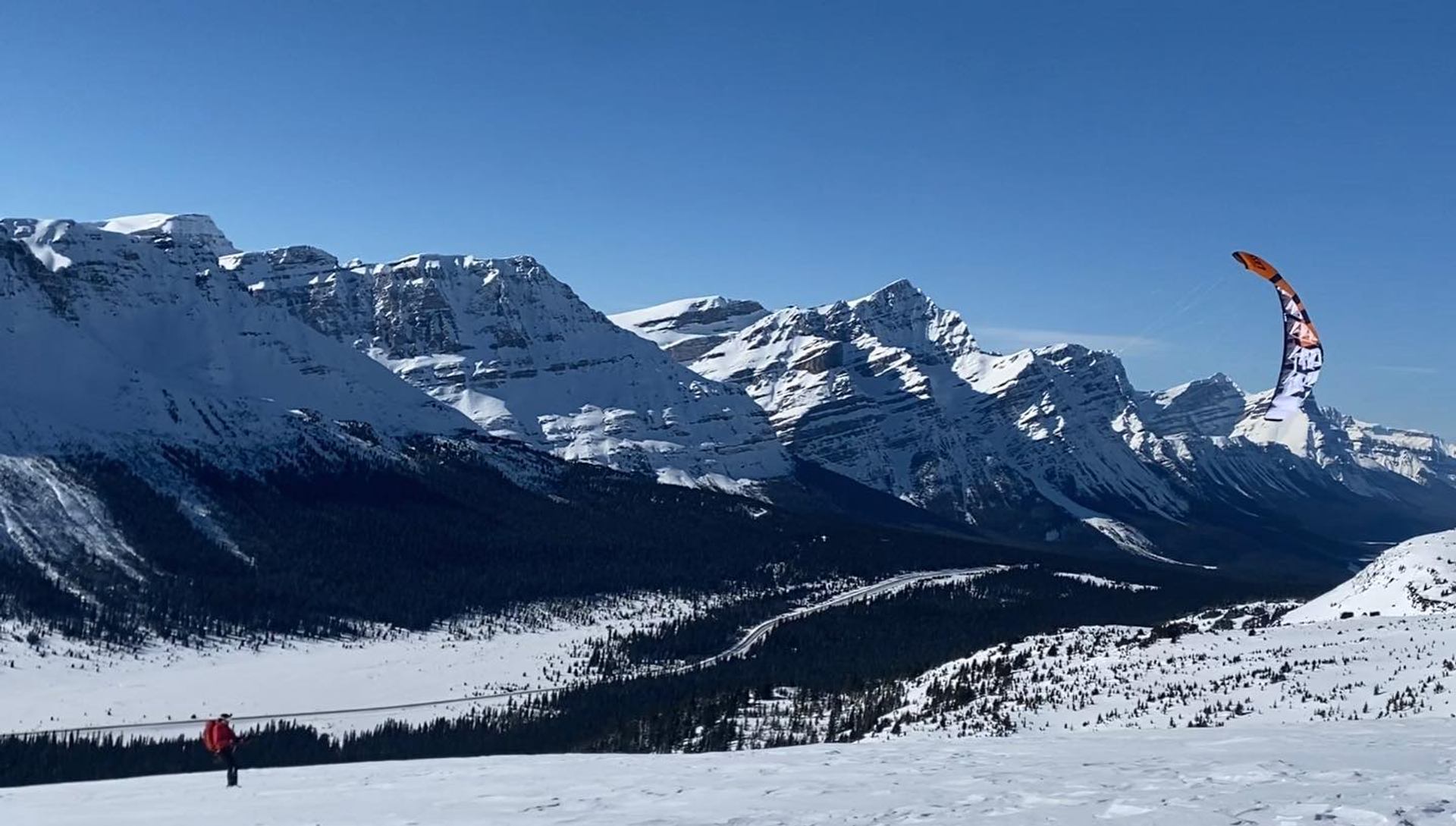 Kite skier travels across a snowy mountain ridge with dramatic peaks and a bright blue sky behind.