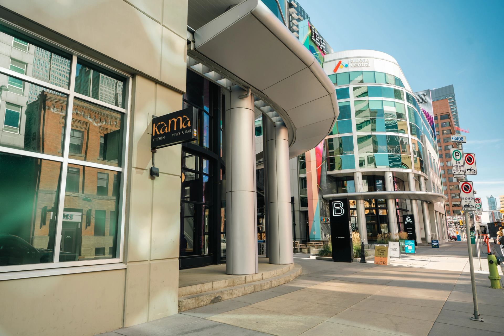 Street view of kama restaurant entrance beside modern glass office building with signs.