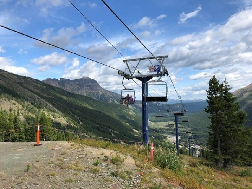 Chairlift carries riders above a forested mountain valley with distant rocky peaks.
