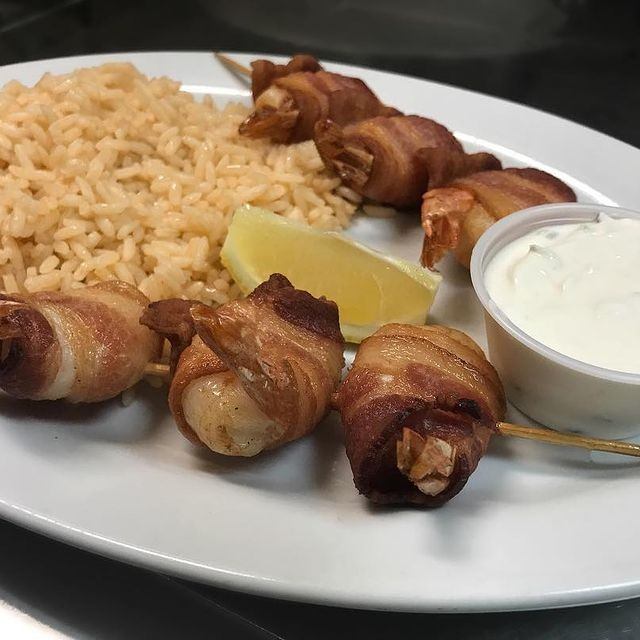 Bacon-wrapped skewers with rice, lemon wedge, and tzatziki sauce.