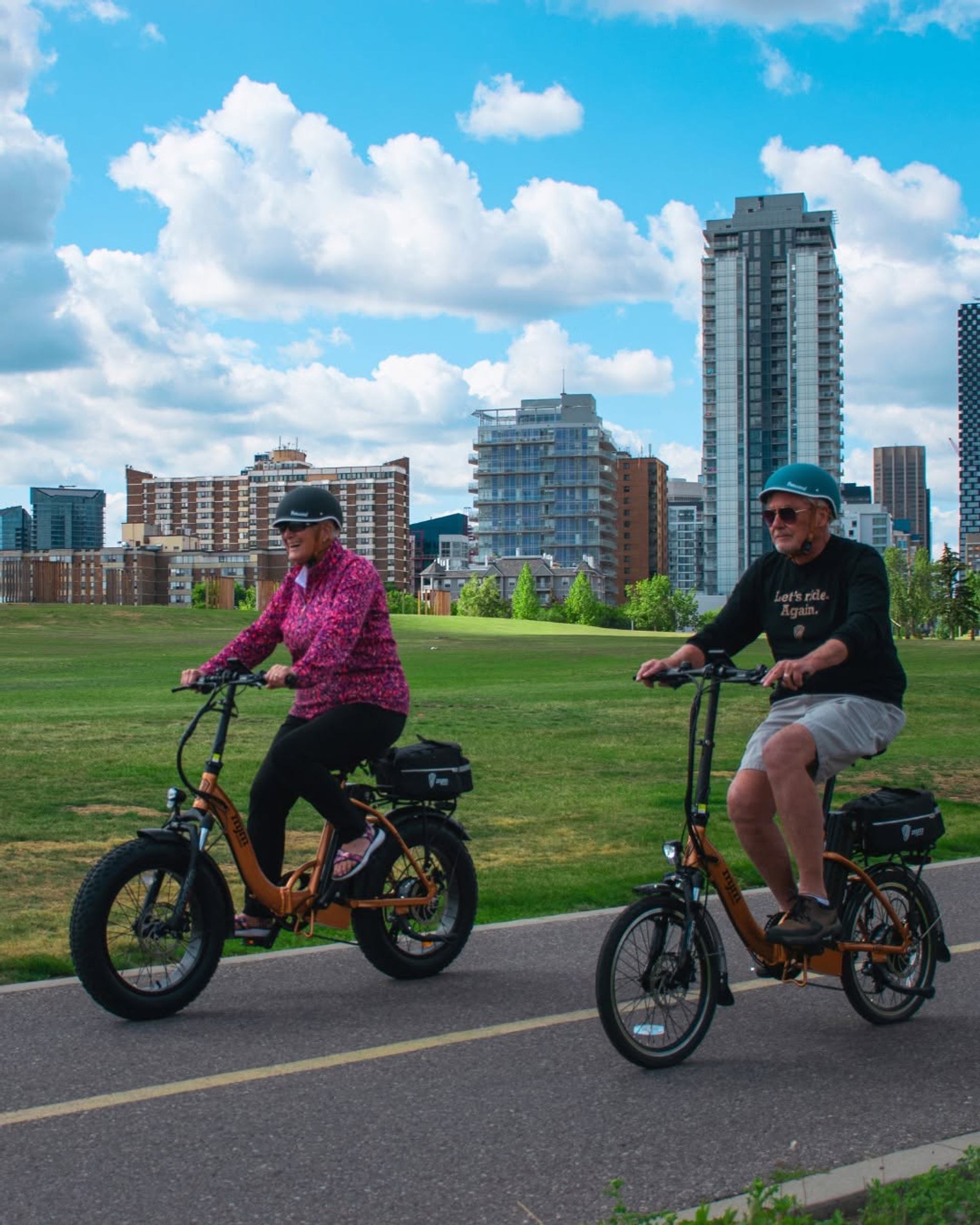 Two people wearing helmets ride electric bicycles along a paved pathway through a green park, with tall buildings and clouds visible in the distance.