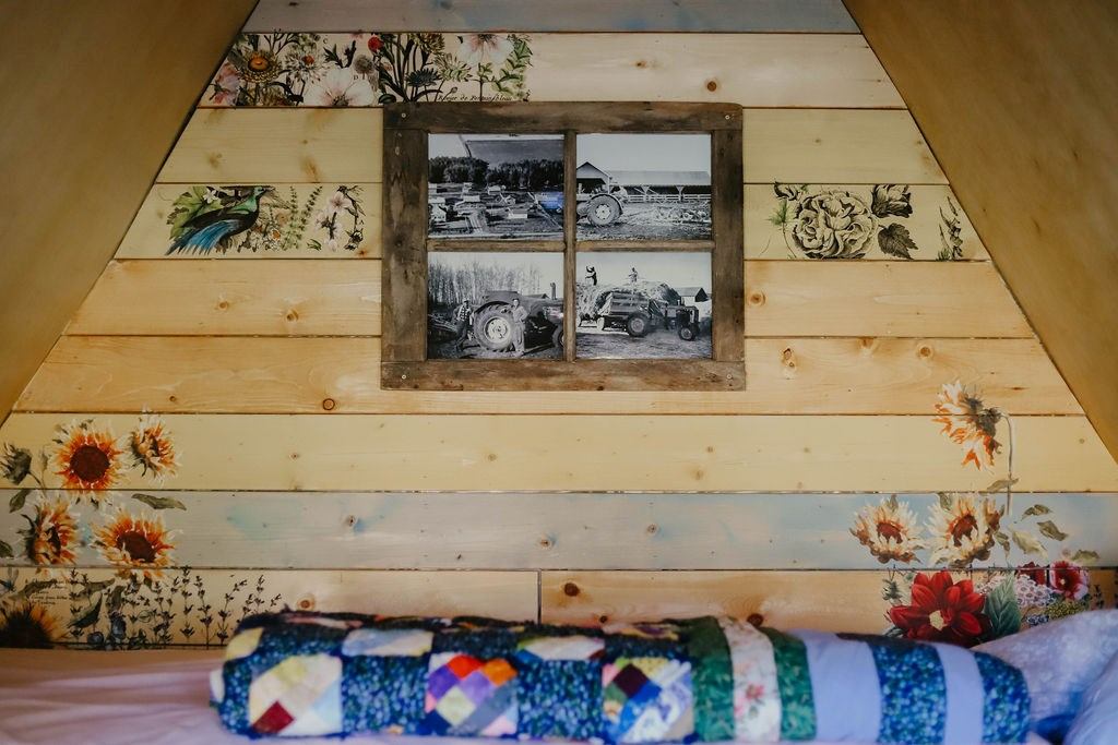The image shows a rustic wooden wall with floral paintings and a wooden-framed window displaying vintage tractor photos. A colorful patchwork quilt is in the foreground, conveying a cozy, nostalgic tone.