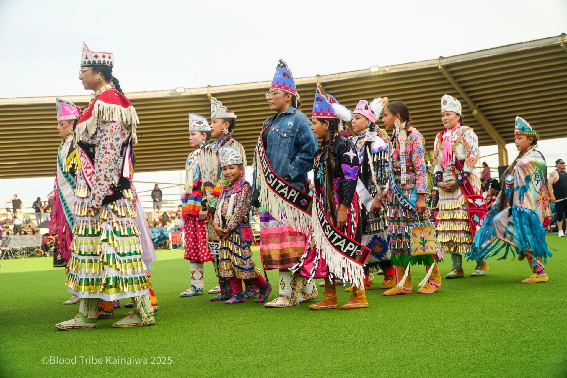 Group of dancers in colourful regalia stand on green field at outdoor event.