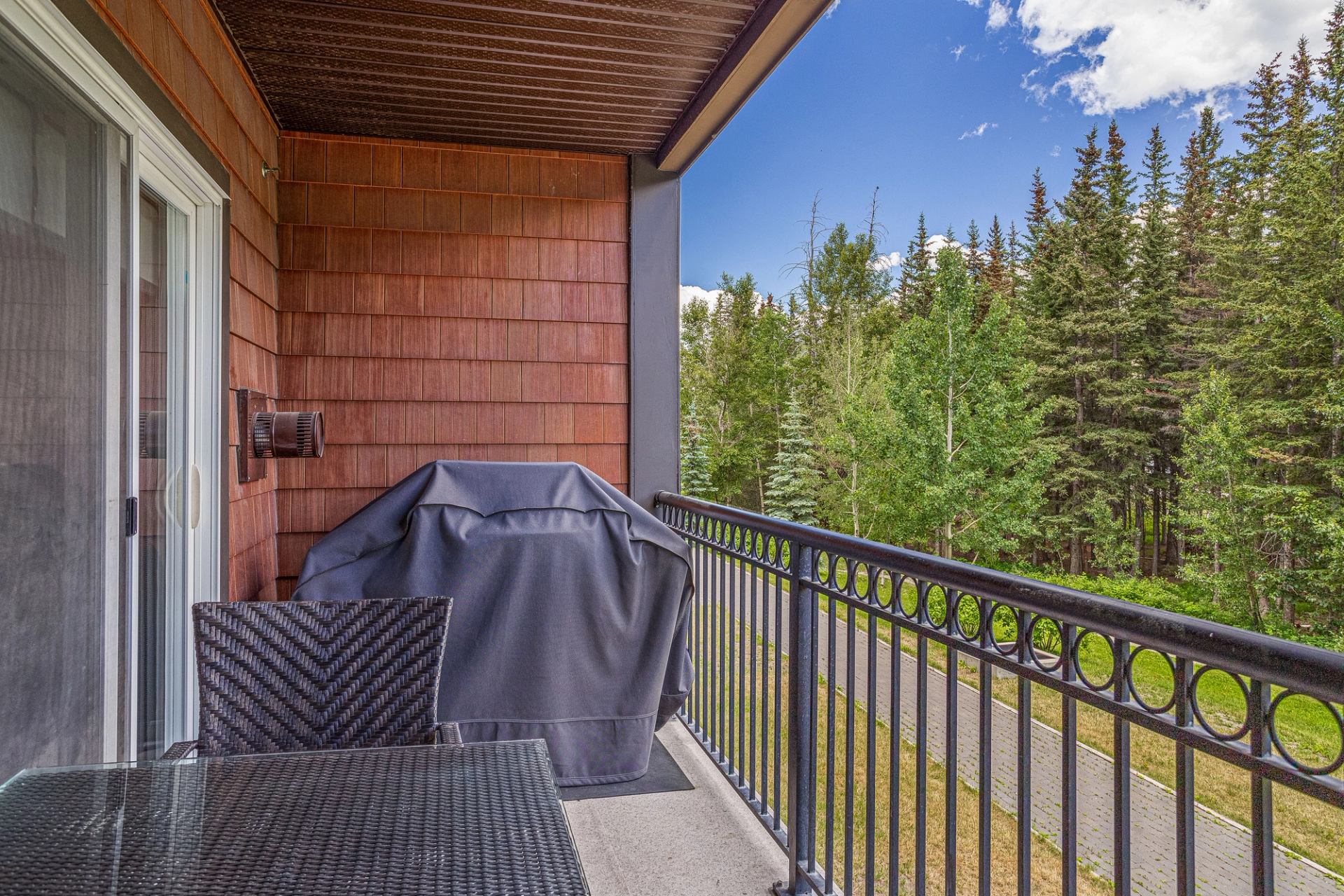 Covered balcony with patio furniture, barbecue and railing overlooking green trees.