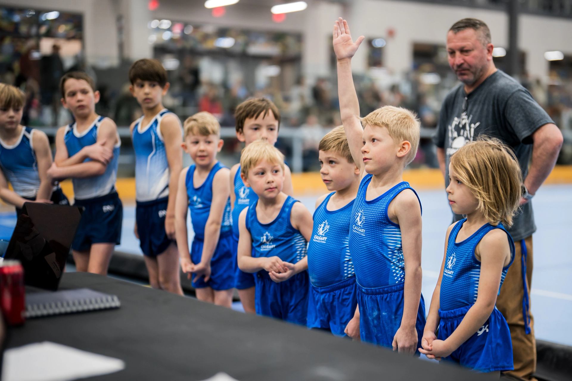 Group of young gymnasts in blue uniforms standing in a line at an indoor event.